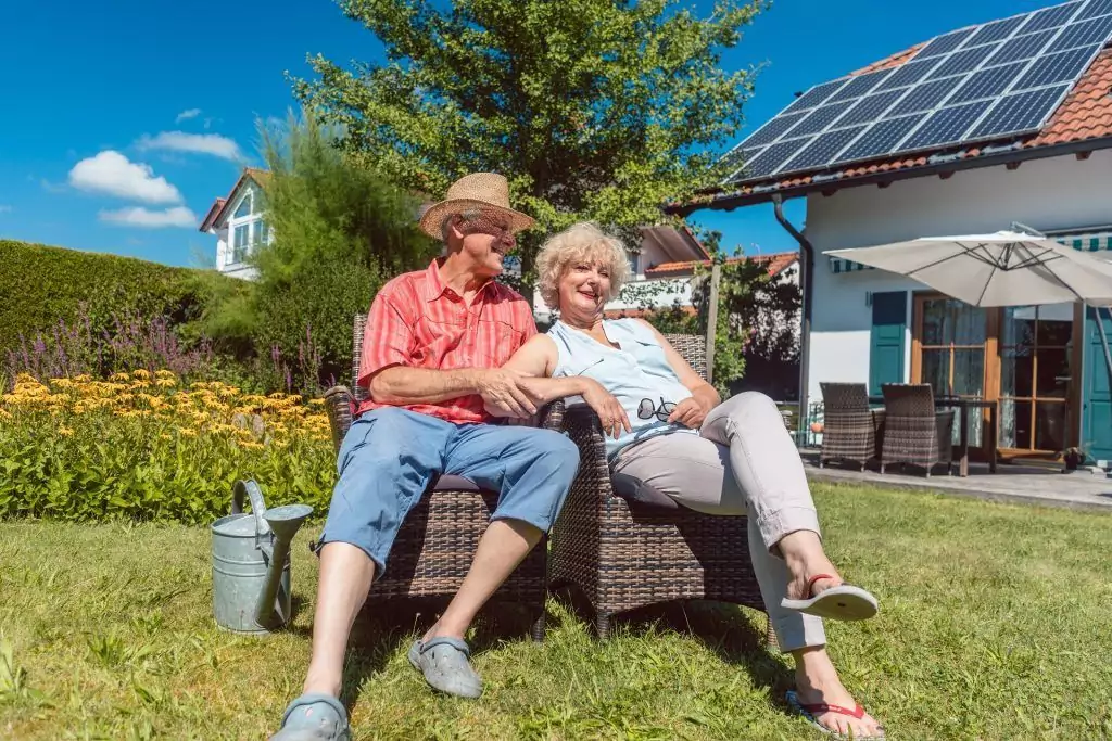 Ehepaar sitzt entspannt im sonnigen Garten und freut sich über erneuerbare Energien in Form einer Photovoltaikanlage auf dem Dach des Hauses